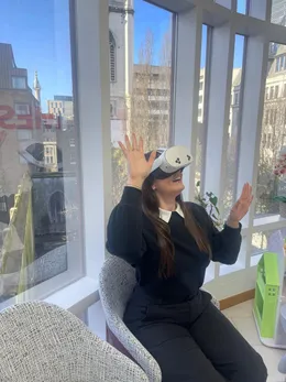 Someone in a headset laughing with both hands raised, in an armchair by a sunlit window.