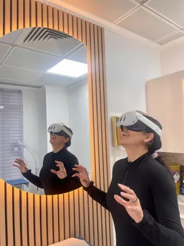 Someone wearing a virtual reality headset, hands raised in front of them, reflected in a mirror set into a wall of pale oak slats.