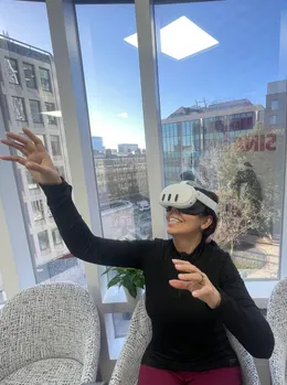 Someone in a headset laughing, one arm stretched above them, with blue sky and rooftops through the window behind.