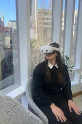 Someone in a headset sitting still with their hands in their lap, turned towards tall windows and a bright sky.
