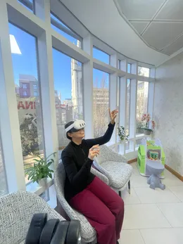 Someone in a headset reaching upward from an armchair, beside tall windows with plants and London rooftops behind.